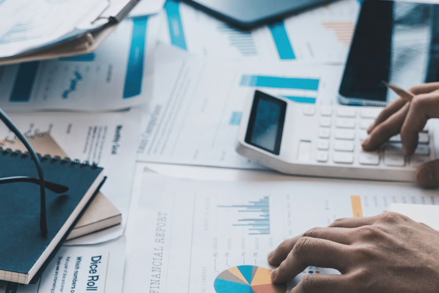 Blog feature image showing a close up of a messy desk with scattered papers; one pair of hands holds a clipboard and another pair of hands use a calculator. It represents the AP and AR teams working hard to fulfil the invoicing process.