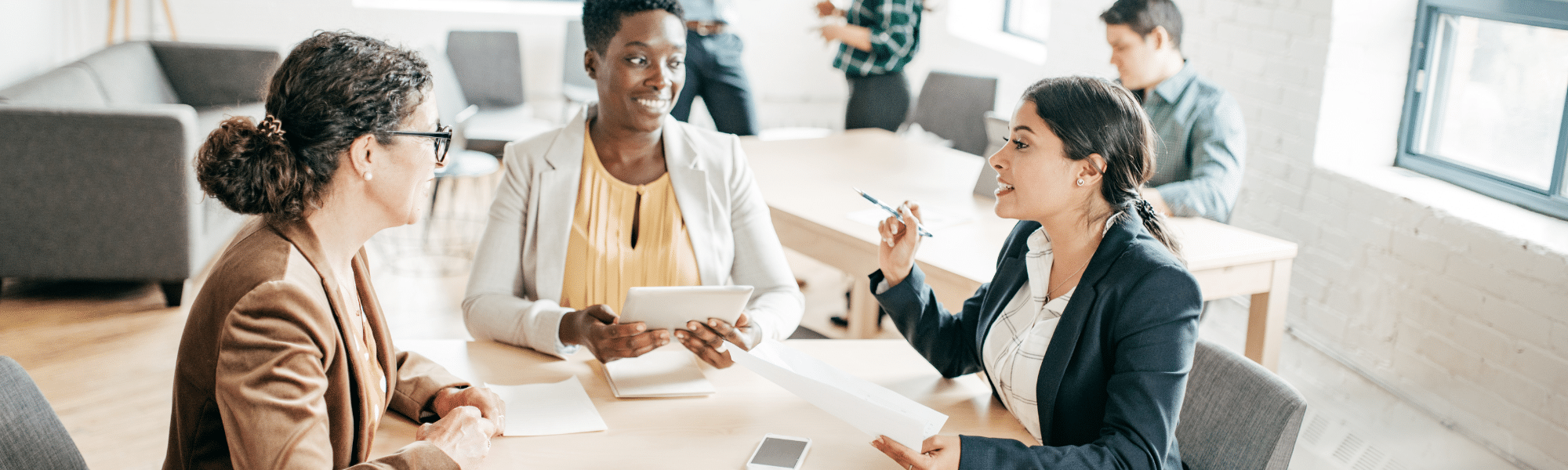 A blog feature image shows three female accountants in an office, discussing work whilst holding electronic gadgets. They sit around a desk filled with invoices, symbolising the shift from paper invoices to electronic invoicing software in the AP and AR team.