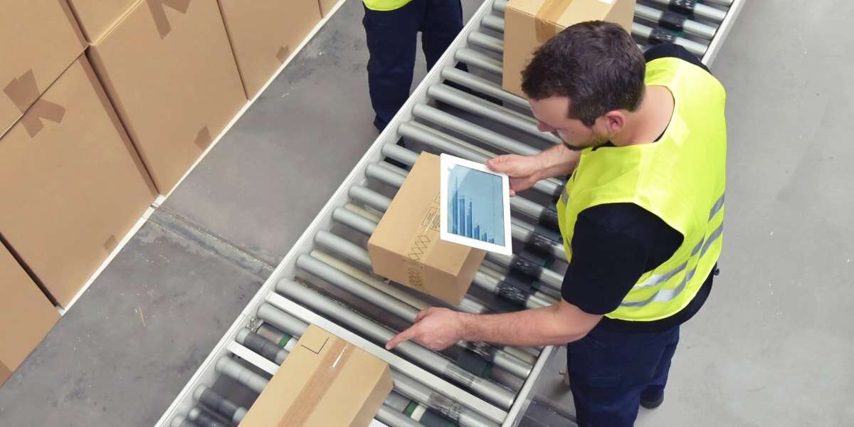 Birdseye view of male workers in warehouse overseeing conveyer belt of packaged goods, with electronic tablets. Representing blog titled 'Expanding the PEPPOL network with new logistics capabilities'