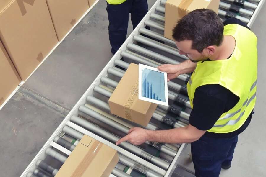 Birdseye view of male workers in warehouse overseeing conveyer belt of packaged goods, with electronic tablets. Representing blog titled 'Expanding the PEPPOL network with new logistics capabilities'