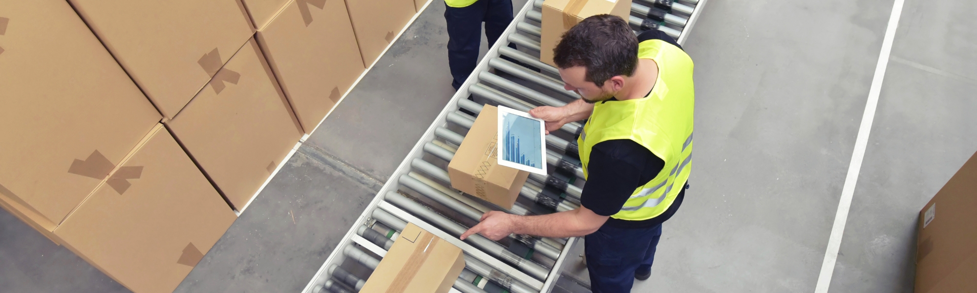 Birdseye view of male workers in warehouse overseeing conveyer belt of packaged goods, with electronic tablets. Representing blog titled 'Expanding the PEPPOL network with new logistics capabilities'