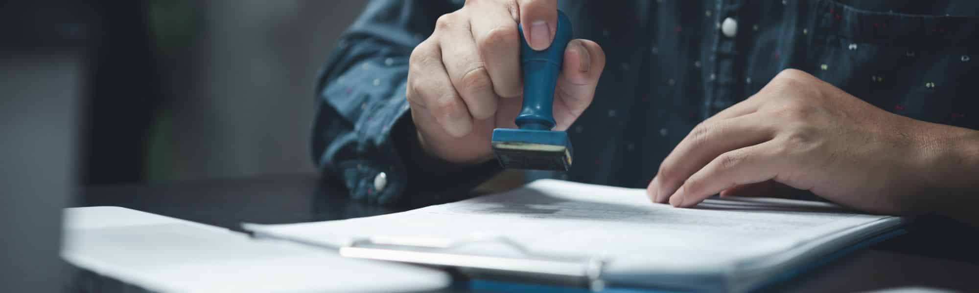 Close up photo of a governmental executive holding an approval stamp ready to sign-off an official document. Representing a blog focused on how businesses can remain on top of tax regulatory compliance